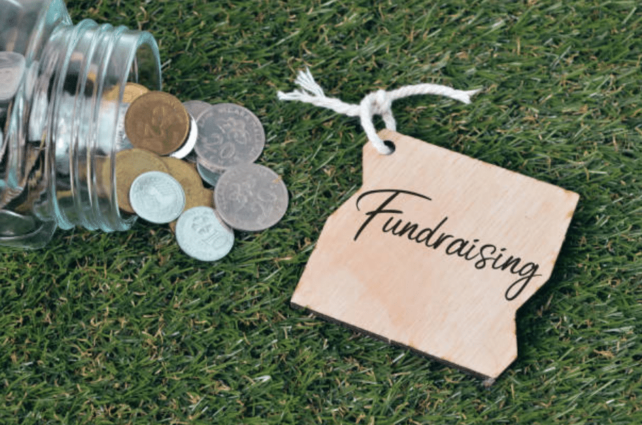 A glass jar tipped over, spilling coins on grass next to a wooden tag labeled 'Fundraising'.